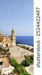 A stunning view of the Chiesa di San Gennaro in Praiano shows its grand plaza with intricate mosaic patterns, set against the backdrop of the sparkling sea. A serene mix of architecture and nature.