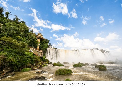 Stunning view of Iguaçu Falls in Brazil, featuring a powerful waterfall, lush greenery, and an observation tower. Blue sky with white clouds complements the natural beauty. - Powered by Shutterstock