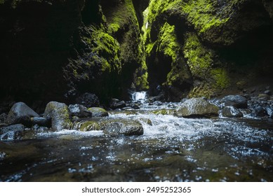 A stunning view of a narrow canyon in Iceland, featuring steep dark walls covered in vibrant green moss and a clear stream flowing over small rocks and pebbles. - Powered by Shutterstock