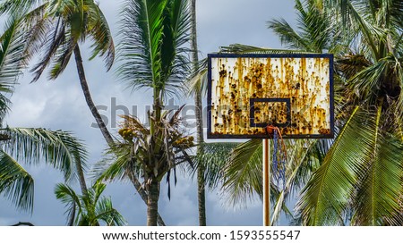 Stunning view of an outdoor basketball court on the island with a tropical background look of palm trees just next to the sea. Abandoned basketball field with rust on the basket board. 