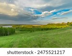 A stunning view of a river valley from a grassy hillside, with lush greenery, fluffy clouds, and a tranquil blue sky. The scene captures the beauty of a spring evening.