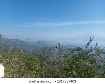 Stunning view of rolling hills in Java, Indonesia, with a distant, misty volcano range under a vast blue sky. Foreground features natural bushes and dry branches.