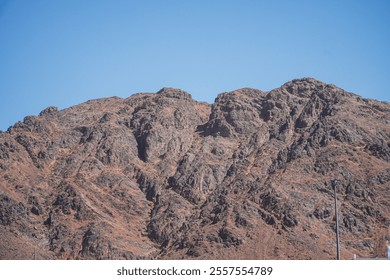 A stunning view of a rugged mountain range against a vibrant blue sky. The texture and colors of the rocks are captivating. Majestic Rocky Uhud Mountain Landscape Under a Clear Blue Sky. - Powered by Shutterstock