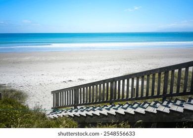 A stunning view of a serene beach featuring wooden steps leading down to the soft sand. The clear blue water and bright sky create a perfect vacation scene. - Powered by Shutterstock