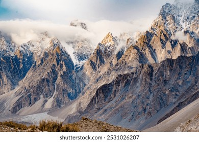 A stunning view of sharp, jagged mountain peaks partially covered by clouds, with golden sunlight casting a warm glow on the rocky cliffs, creating a dramatic and awe-inspiring landscape.