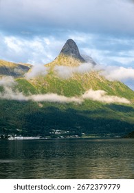A stunning view of a sharp mountain peak in Norway partially shrouded in clouds, rising above a tranquil lake | Lush green slopes meet the water's edge, with a small settlement visible below.