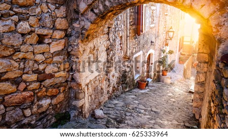 Stunning view of the street in old town of Ulcinj on sunset. Pots with flowers standing on paved pavement along stone wall. The sun light on stone arch. Travel Montenegro