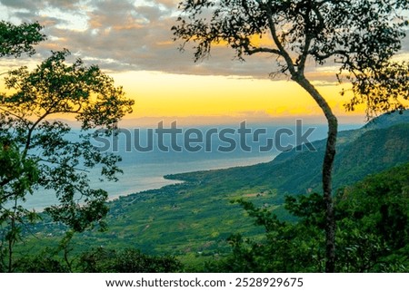 A stunning view of the sun setting over a coastal landscape in Malawi, framed by lush green trees and overlooking the vast expanse of Lake Malawi, with vibrant colors in the sky.