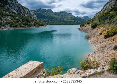 A stunning view of a turquoise lake surrounded by rocky mountains and lush greenery under a cloudy sky. - Powered by Shutterstock