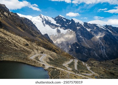 A stunning winding road snakes through the rugged Peruvian Andes, framed by towering snow-capped mountains and a serene mountain lake under a bright, blue sky.
 - Powered by Shutterstock