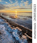 Stunning winter sunset scene with a frozen beach and icy shore covered in snow and ice. Fiery orange sun setting over the calm Baltic Sea, creating a serene and beautiful landscape.