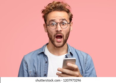 Stupefied Caucasian guy with surprised expression, holds cellular in hands, recieves unexpected notification, wears round spectacles, keeps mouth opened, isolated over pink studio background