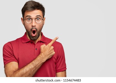 Stupefied emotional bearded handsome male with jaw dropped points aside at upper right corner, wears red clothing, isolated over white background with blank space for your advertising content