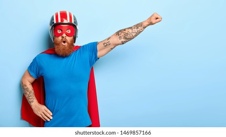 Stupefied emotive man with ginger beard being cartoon character, keeps arm in flying gesture, wears protective headgear, blue t shirt and red cloak, has shocked expression, saves our universe