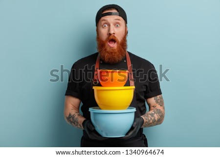 Stupefied emotive tattooed chef holds three plastic dishes, opens mouth widely, has mauch work at kitchen, has red beard, isolated over blue background. Surprised man with colorful pots indoor