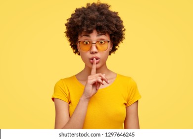 Stupefied secret dark skinned woman makes gesture quietly, asks remain silent, gossips about something, looks mysteriously as tells secret, wears bright yellow t shirt, stands indoor. Shh, dont speak