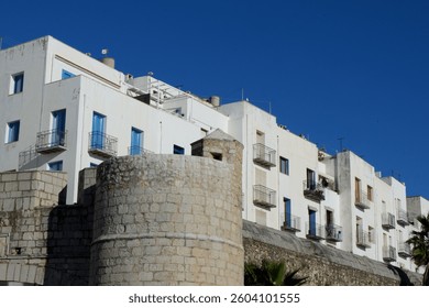 A sturdy stone wall stands adjacent to contemporary white apartment buildings, showcasing a blend of historical and modern architecture under a vibrant blue sky.