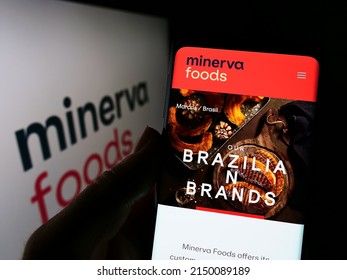 Stuttgart, Germany - 03-30-2022: Person holding mobile phone with webpage of Brazilian foods company Minerva SA on screen in front of logo. Focus on center of phone display. Unmodified photo.