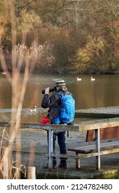 Stuttgart, Germany - January 06, 2022: Male Wildlife Photographer Watching The Water Birds At The Lake. Man With System Camera On The Shore With Backpack In Winter.