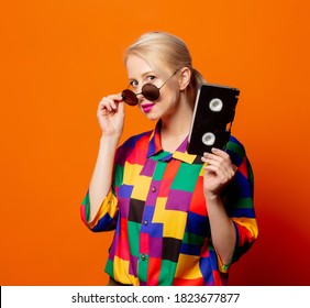 Style Blonde In 90s Clothes With VHS Cassette On Orange Background