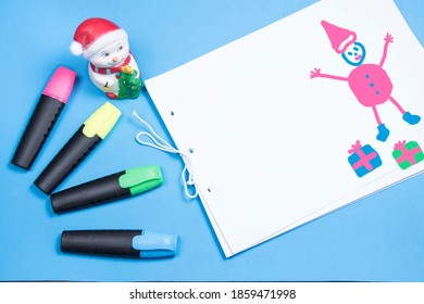 Children’s Style Drawing Christmas. Album, Red, Blue, Yellow And Green Felt Tip Pens, Toy Snowman. Child's Drawing Santa Claus. Copyspace For Text. Blue Background. 