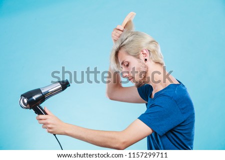 Style and fashion. Young trendy male hairstylist barber with new idea of look changing. Blonde man holding hair dryer and comb creating new hairdo, on blue