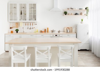 Style Minimalism. Stylish Kitchen In White And Brown Wood. Dining Table, Utensils And Kitchenware On Shelves, Small White Refrigerator Near Window