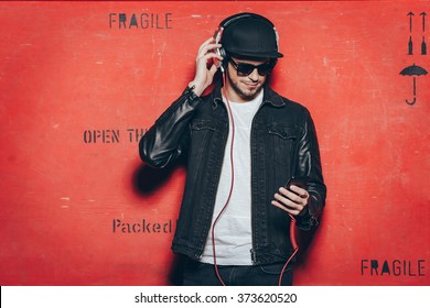 Style and music. Handsome young man adjusting his headphones and looking at his smart phone while standing against red background - Powered by Shutterstock