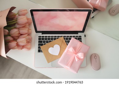 Styled feminine desk workspace with pink tulips, laptop computer and envelope. Top view notebook screen. Pink gift box and pink computer mouse.