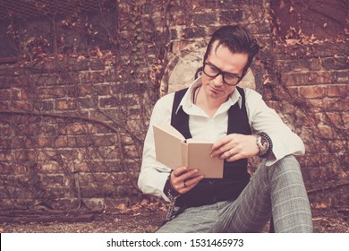Styled man reading book while sitting outdoors against brick wall. 