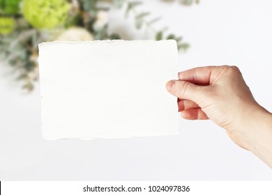 Styled Stock Photo. Feminine Wedding, Birthday Greeting Card Mockup Scene With Woman's Hand In Holding Blank Paper Card. Blurred Floral Background. Empty Space. Picture For Blog Or Social Media.