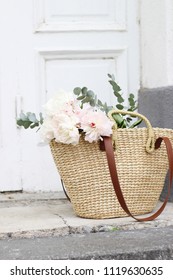 Styled stock photo. Feminine wedding still life composition with straw French basket bag with pink peonies flowers and eucalyptus bouquet. Old white door in the background. Vertical composition.