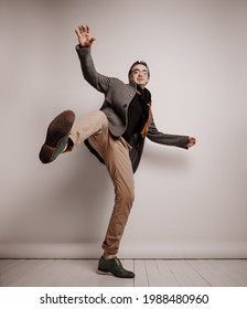 Stylish Adult Man Hipster In Pants And Plaid Jacket Stands Holding Foot Up, Making Giant Step, Has Lost Balance, Falling Down Waving Hands Over Grey Background