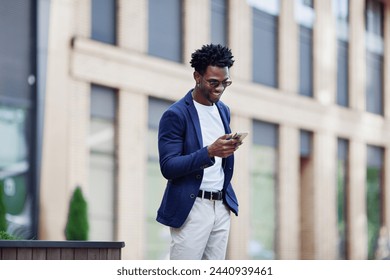 Stylish African American man in smart casual clothes, with mobile phone against backdrop of city. Fashionable guy with smartphone outdoors. - Powered by Shutterstock