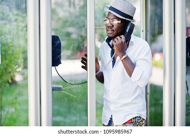 Stylish African American Man In White Shirt And Colored Pants With Hat And Glasses Posed Outdoor At Telephone Booth. 