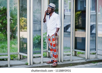 Stylish African American Man In White Shirt And Colored Pants With Hat And Glasses Posed Outdoor At Telephone Booth. Black Fashionable Model Boy.