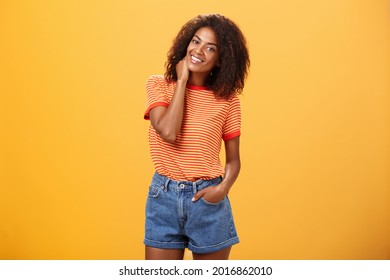 Stylish African American slim woman with curly medium hair holding hand in denim shorts tilting head, rubbing neck while smiling friendly feeling uncomfortable or shy in new company over orange wall