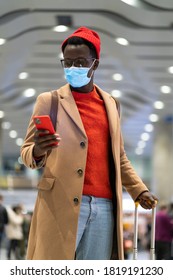 Stylish African American traveler man in beige coat, using phone and calls taxi, stands in airport terminal, wear face medical mask to protect yourself from contact with flu virus, pandemic covid-19