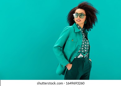 stylish african american woman in jacket with hands in pocket looking away isolated on turquoise