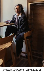 Stylish African American Woman In Oversize Suit Holding Smartphone While Sitting In Cafe