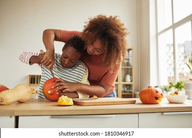Stylish African American woman spending time with her son standing at table carving pumpkin for Halloween with kitchen knife together