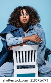 Stylish African American Woman With Wavy Hair Sitting On Chair With Crossed Arms Isolated On Blue