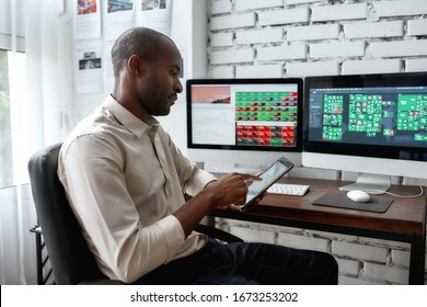 Stylish African Businessman, Trader Sitting By Desk In Front Of Multiple Monitors And Using His Smartphone To Check Charts While Working In The Office. Stock Trading, People Concept.