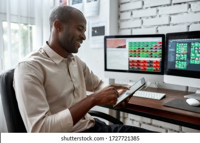 Stylish African Businessman, Trader Sitting By Desk In Front Of Multiple Monitors And Using His Smartphone To Check Charts While Working In The Office. Stock Trading, People Concept.