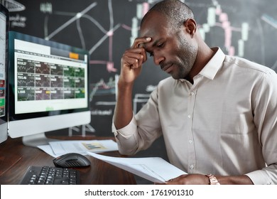 Stylish African Businessman, Trader Sitting By Desk In Front Of Multiple Monitors. He Is Getting Tired While Looking Through Papers. Stock Trading, People Concept.