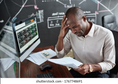 Stylish African Businessman, Trader Sitting By Desk In Front Of Multiple Monitors. He Is Getting Angry While Looking Through Papers. Stock Trading, People Concept.