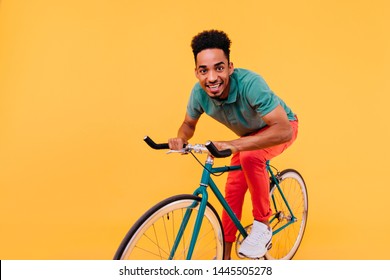 Stylish African Guy In Green T-shirt And White Sneakers Posing On Bicycle. Studio Shot Of Blissful Black Man Riding On Bike On Yellow Background.