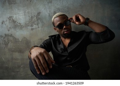 Stylish African Handsome Male Model Wearing Black Silk Shirt, Trousers And Sunglasses, Sitting Leaning On Grey Concrete Wall