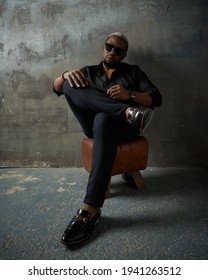 Stylish African Handsome Male Model Wearing Black Silk Shirt, Trousers And Sunglasses, Sitting Leaning On Grey Concrete Wall