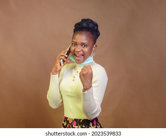 Stylish African Lady Wearing Nose Mask, And Turtle Neck Top As She Happily Makes Call With The Smart Phone In Her Hands